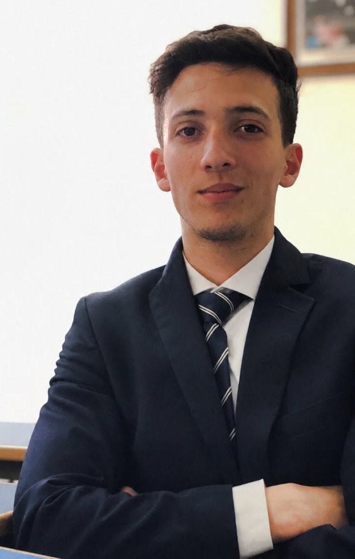 The founder of NŪR Concierge, arms crossed, seated at a desk in a navy suit and striped tie, looking directly at the camera.